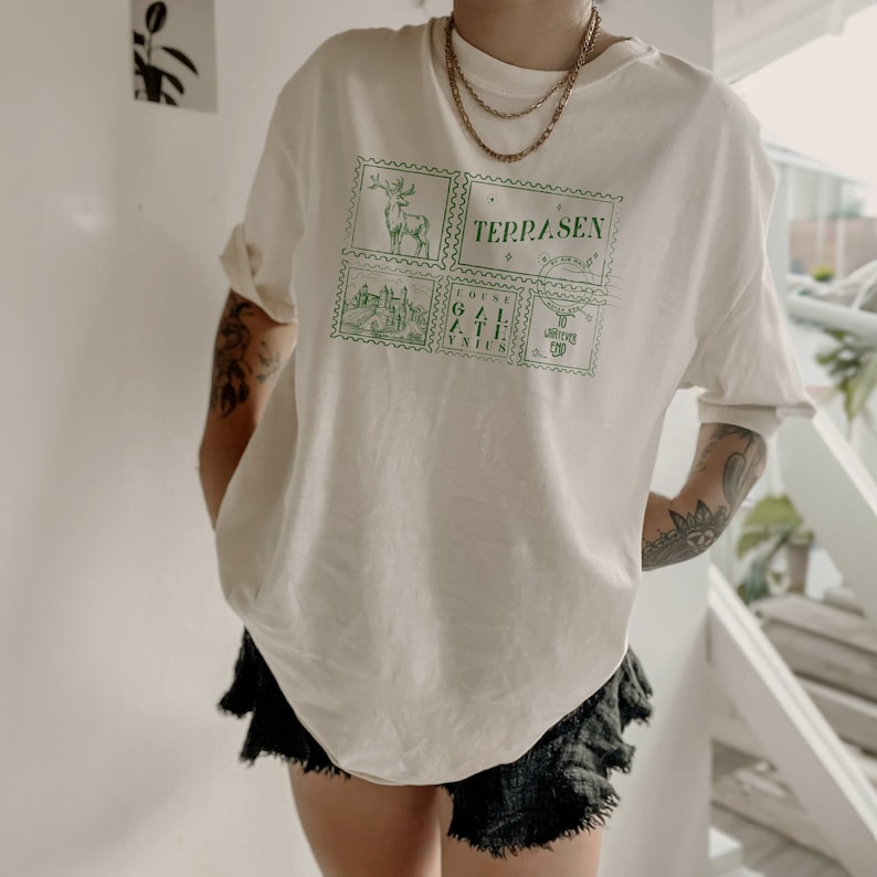 May include: A white t-shirt with a green graphic print of stamps featuring a deer, a building, and the text "TERRASEN", "COFFEE GATE", and "TO WHATEVER END".
