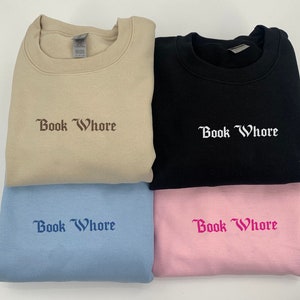 Puede incluir: Cuatro sudaderas de diferentes colores con el texto "Book Whore" bordado en una fuente g&oacute;tica. Las sudaderas son beige, negra, azul claro y rosa.
