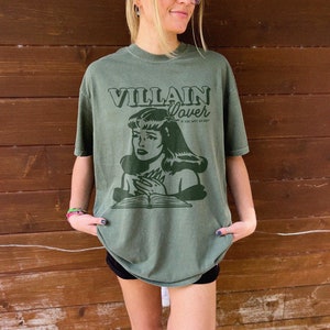 Pode incluir: Uma camiseta verde com um gr&aacute;fico de estilo vintage de uma mulher lendo um livro. O texto na camiseta diz "VILLAIN Lover" e "IF I SAY, WHY SO HOT?!".