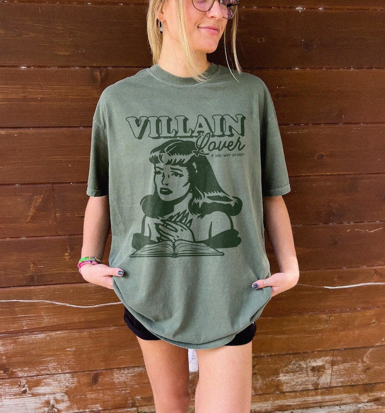 Pode incluir: Uma camiseta verde com um gr&aacute;fico de estilo vintage de uma mulher lendo um livro. O texto na camiseta diz "VILLAIN Lover" e "IF I SAY, WHY SO HOT?!".