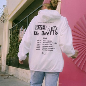 Peut inclure: Un sweat &agrave; capuche blanc avec le texte "ENEMIES TO LOVERS" et une liste de dates et de phrases. Le sweat &agrave; capuche a une capuche &agrave; cordon et des manches longues. La personne porte une casquette rose et un jean bleu clair. L'arri&egrave;re-plan comprend une porte rose et un b&acirc;timent.