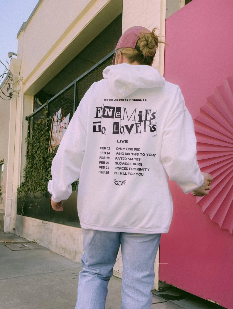 Peut inclure: Un sweat &agrave; capuche blanc avec le texte "ENEMIES TO LOVERS" et une liste de dates et de phrases. Le sweat &agrave; capuche a une capuche &agrave; cordon et des manches longues. La personne porte une casquette rose et un jean bleu clair. L'arri&egrave;re-plan comprend une porte rose et un b&acirc;timent.