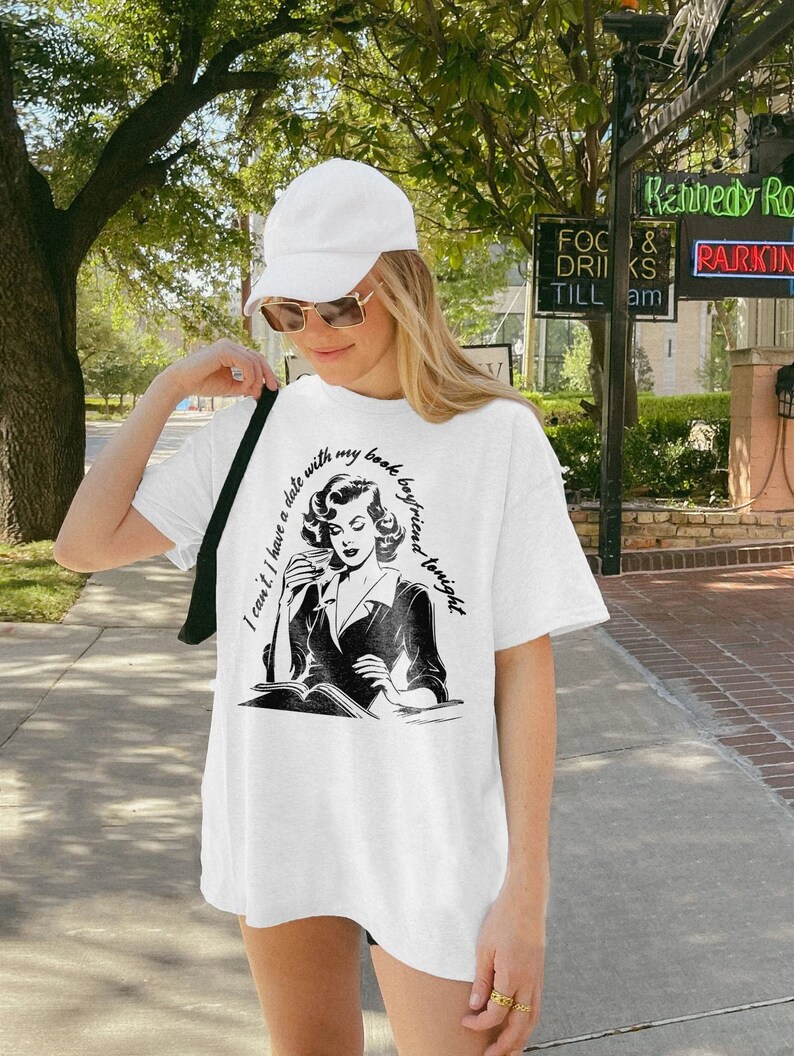 May include: White oversized t-shirt with a black and white illustration of a woman reading a book and drinking from a cup. The text on the shirt reads "I can't. I have a date with my book boyfriend tonight."