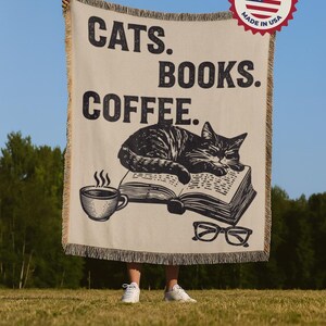 May include: A beige throw blanket with the words "CATS. BOOKS. COFFEE." in black. A graphic of a cat sleeping on a book, a coffee cup, and glasses are also featured. The blanket has a fringed edge and a "Proudly Made in USA" sticker.