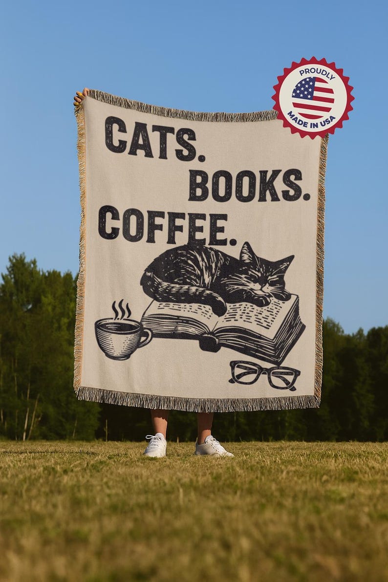 May include: A beige throw blanket with the words "CATS. BOOKS. COFFEE." in black. A graphic of a cat sleeping on a book, a coffee cup, and glasses are also featured. The blanket has a fringed edge and a "Proudly Made in USA" sticker.