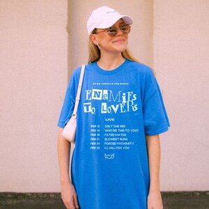 May include: A blue t-shirt with the text "BOOK ADDICTS PRESENTS ENEMIES TO LOVERS LIVE" and a list of dates and events. The shirt has a white graphic of a heart with wings.