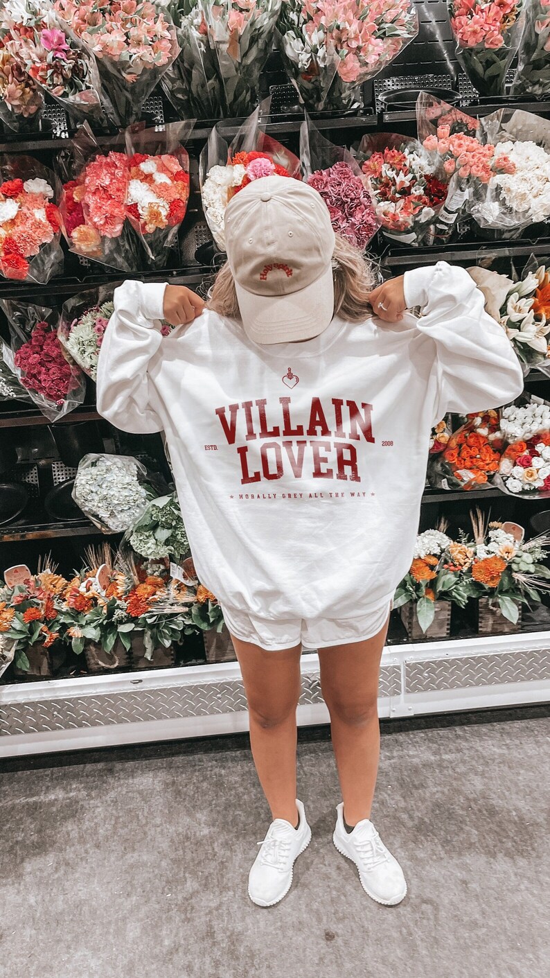 May include: White sweatshirt with red text that reads "VILLAIN LOVER" with the years 1978 and 2008 on either side of the text. The text also includes "MORALLY GREY ALL THE WAY" in smaller font. A person is wearing the sweatshirt with a beige baseball cap that says "Holy Water" on it.