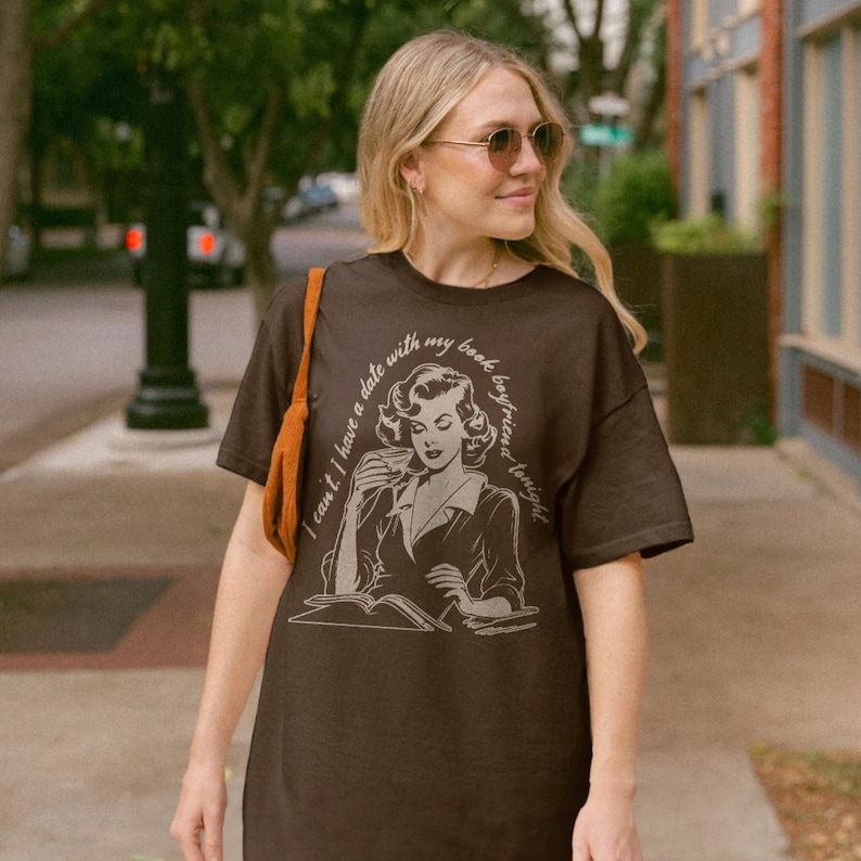 May include: A brown t-shirt with a vintage illustration of a woman reading a book and drinking from a cup. The text on the shirt reads "I can't I have a date with my book boyfriend tonight."