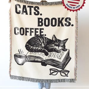 May include: Cream-colored throw blanket with black text reading "CATS. BOOKS. COFFEE." A black and white illustration of a cat sleeping on a book, a coffee cup, and glasses are also featured. The blanket has a fringed edge.