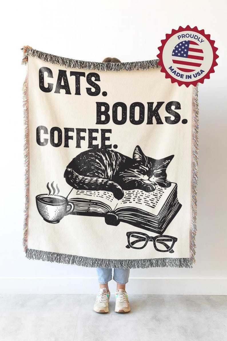 May include: Cream-colored throw blanket with black text reading "CATS. BOOKS. COFFEE." A black and white illustration of a cat sleeping on a book, a coffee cup, and glasses are also featured. The blanket has a fringed edge.