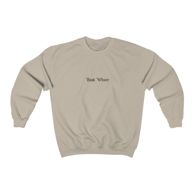 Puede incluir: Una sudadera de color beige claro con el texto "Book Whore" impreso en marr&oacute;n en la parte delantera.