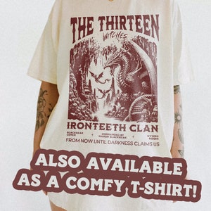 Pu&ograve; includere: Una t-shirt bianca con una stampa grafica bordeaux di un drago e il testo "The Thirteen Witches Ironteeth Clan From Now Until Darkness Claims Us" sul davanti. Il testo "Also Available As A Comfy T-Shirt!" &egrave; stampato in un grande font sotto la grafica.