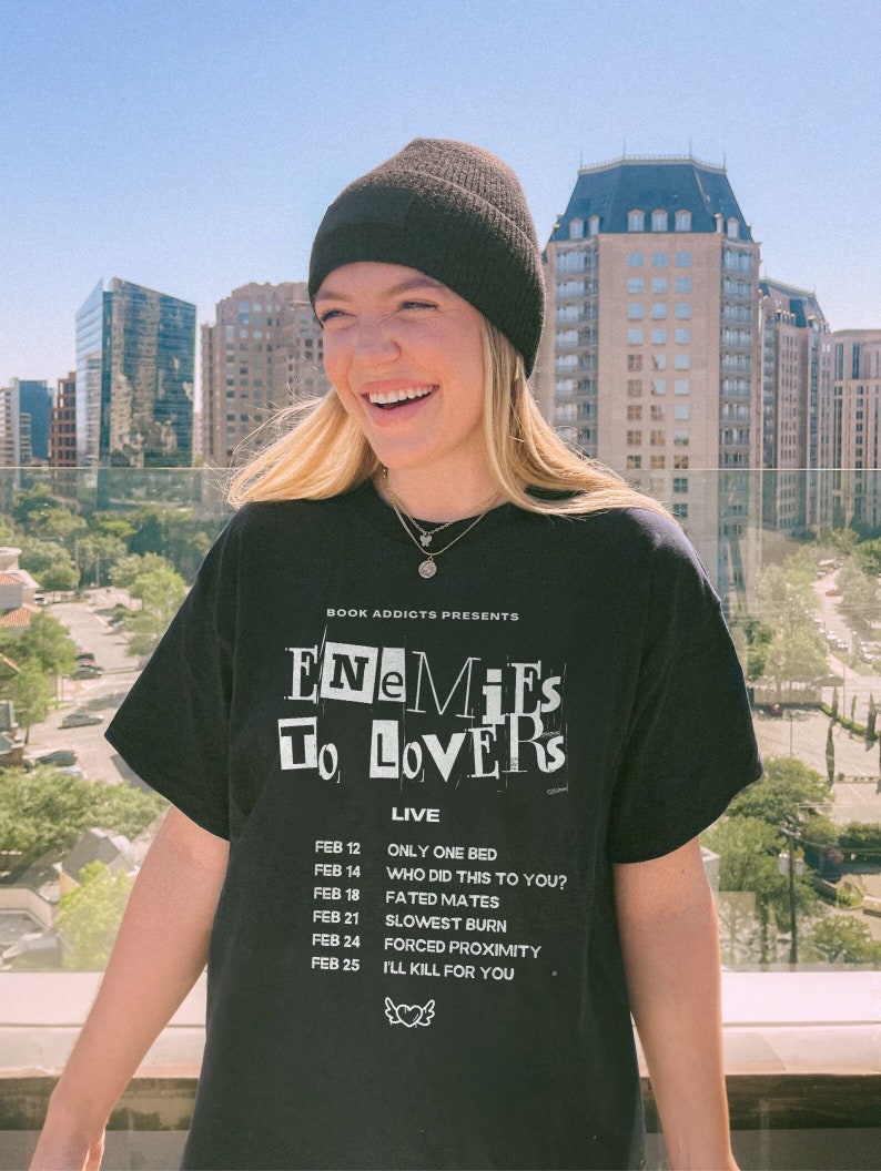 May include: A person wearing a black beanie and a black t-shirt with white text that reads "BOOK ADDICTS PRESENTS ENEMIES TO LOVERS LIVE" and a list of dates and events.