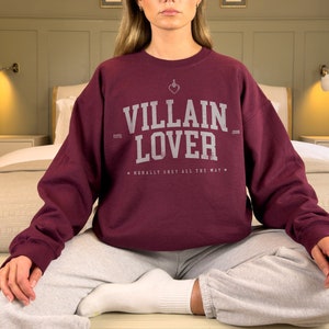 May include: A maroon sweatshirt with the text "Villain Lover" in white, with a small heart and dagger graphic above the text. The text "Morally Grey All The Way" is printed below the main text.