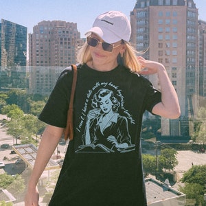 May include: A black t-shirt with a graphic of a woman reading a book and drinking coffee. The text on the shirt reads "I can't I have a date with my book boyfriend tonight".
