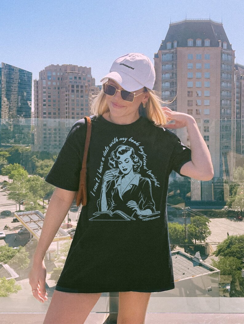 May include: A black t-shirt with a graphic of a woman reading a book and drinking coffee. The text on the shirt reads "I can't I have a date with my book boyfriend tonight".