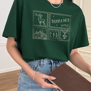 May include: Green short-sleeved t-shirt with a graphic print of four postage stamps featuring a deer, a castle, and the word "TERRASEN".
