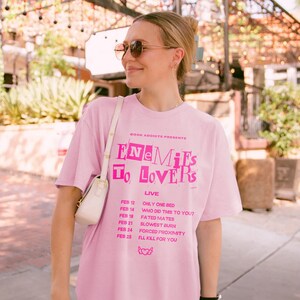 May include: Pink t-shirt with the text "Book Addicts Presents Enemies To Lovers Live" and a list of dates and events printed on the front.