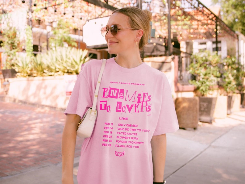 May include: Pink t-shirt with the text "Book Addicts Presents Enemies To Lovers Live" and a list of dates and events printed on the front.