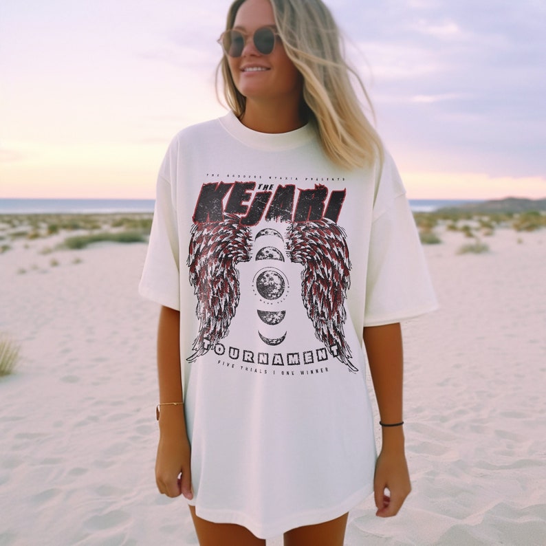 May include: A white oversized t-shirt with a graphic print of a red and black winged design with a moon and stars. The text on the shirt reads "The Kejari Tournament Five Trials One Winner".