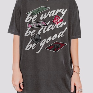 Puede incluir: Una camiseta gris oscuro con un estampado gr&aacute;fico de estilo vintage que dice "be wary be clever be good". El gr&aacute;fico presenta una serie de im&aacute;genes en forma de diamante con varias ilustraciones.