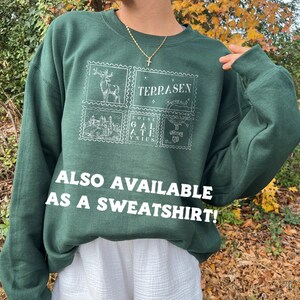 May include: A dark green sweatshirt with a white graphic print of four postage stamps. The stamps feature a deer, a castle, a text design, and a design with the words "To Wherever You Go". The text "Terrasen" is printed on the sweatshirt. The text "ALSO AVAILABLE AS A SWEATSHIRT!" is printed below the graphic.