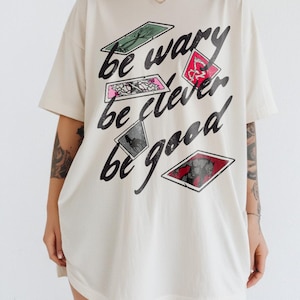 Puede incluir: Una camiseta blanca de gran tama&ntilde;o con un estampado gr&aacute;fico de cartas de juego con el texto "be wary be clever be good".