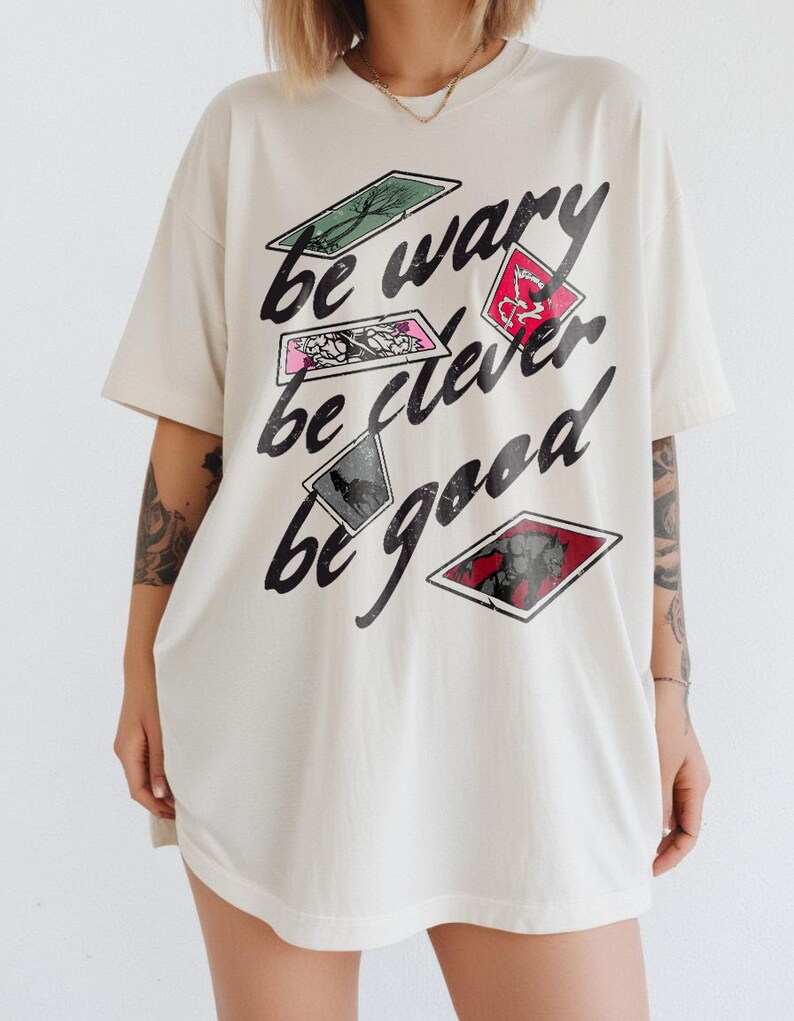 Puede incluir: Una camiseta blanca de gran tama&ntilde;o con un estampado gr&aacute;fico de cartas de juego con el texto "be wary be clever be good".