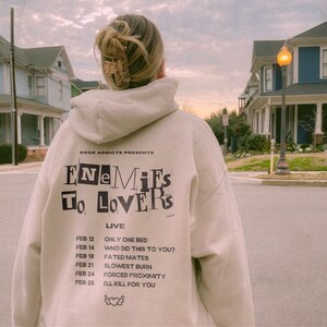Peut inclure: Un sweat-shirt beige avec l'inscription noire "ENEMIES TO LOVERS" et une liste de dates et de titres. Le sweat-shirt est port&eacute; par une personne dans une rue, avec des maisons en arri&egrave;re-plan.