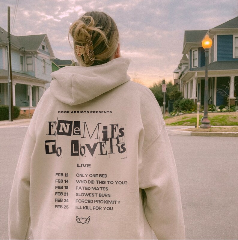 Peut inclure: Un sweat-shirt beige avec l'inscription noire "ENEMIES TO LOVERS" et une liste de dates et de titres. Le sweat-shirt est port&eacute; par une personne dans une rue, avec des maisons en arri&egrave;re-plan.