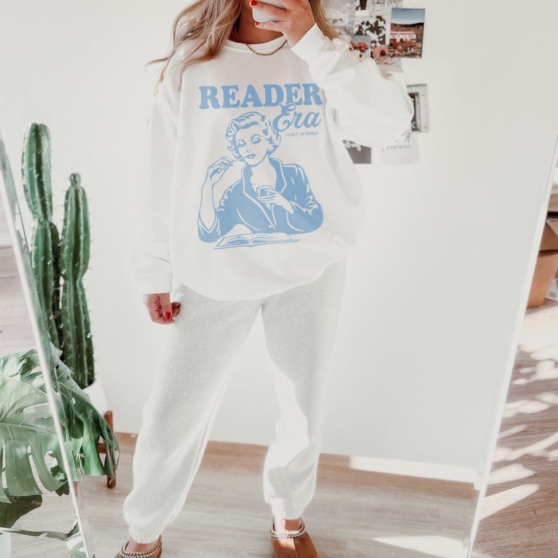 May include: White sweatshirt with a blue graphic of a woman reading a book and the text "Reader Era". The sweatshirt is paired with matching white sweatpants.