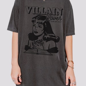 Pode incluir: Uma camiseta cinza escura de tamanho grande com uma estampa gr&aacute;fica preta de uma mulher com um livro e o texto "VILLAIN Lover # BAD. WHY SO HOT?"