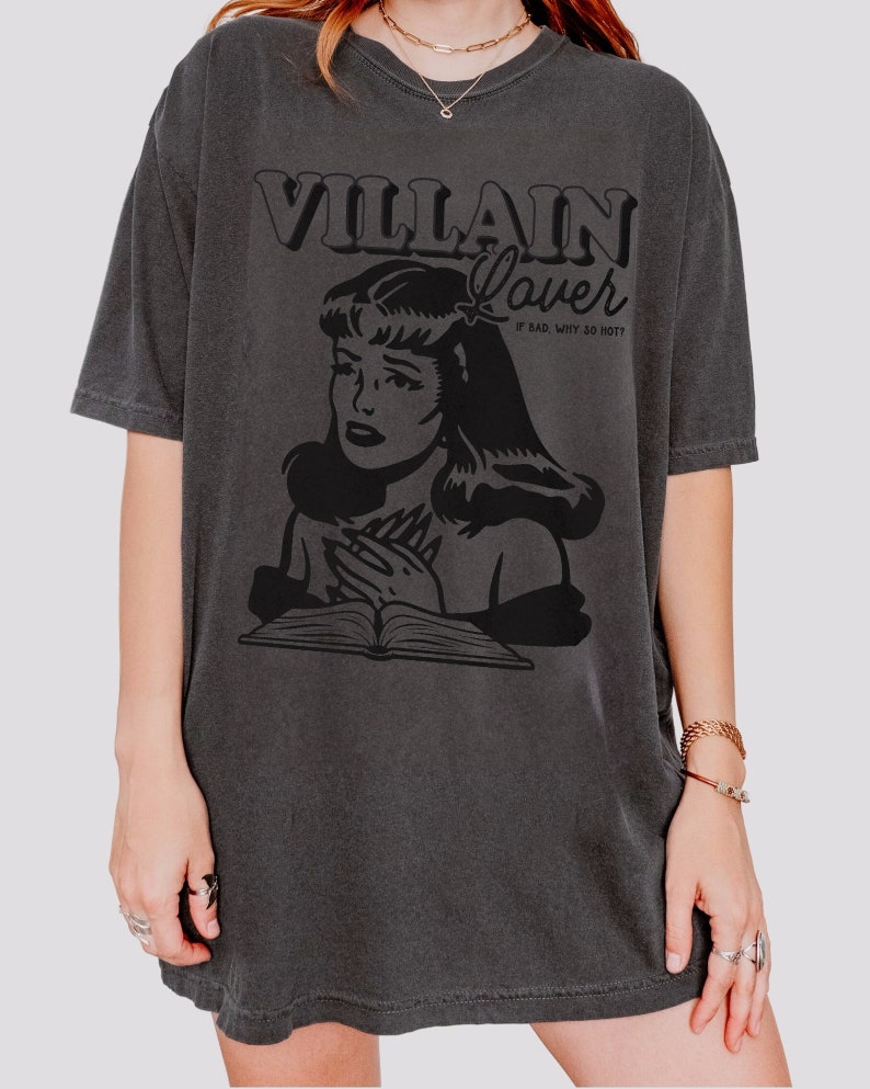 Pode incluir: Uma camiseta cinza escura de tamanho grande com uma estampa gr&aacute;fica preta de uma mulher com um livro e o texto "VILLAIN Lover # BAD. WHY SO HOT?"