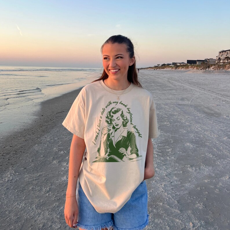 May include: A woman wearing a beige t-shirt with a green graphic of a woman reading a book and the text "I can't have a date with my book boyfriend tonight". She is standing on a sandy beach with the ocean in the background.