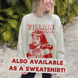 Pode incluir: Uma camisa de moletom cinza claro com uma estampa gr&aacute;fica vermelha de uma mulher com o texto "VILLAIN Lover IF BAD, WHY SO HOT?" e o texto "ALSO AVAILABLE AS A SWEATSHIRT!" em um adesivo branco.