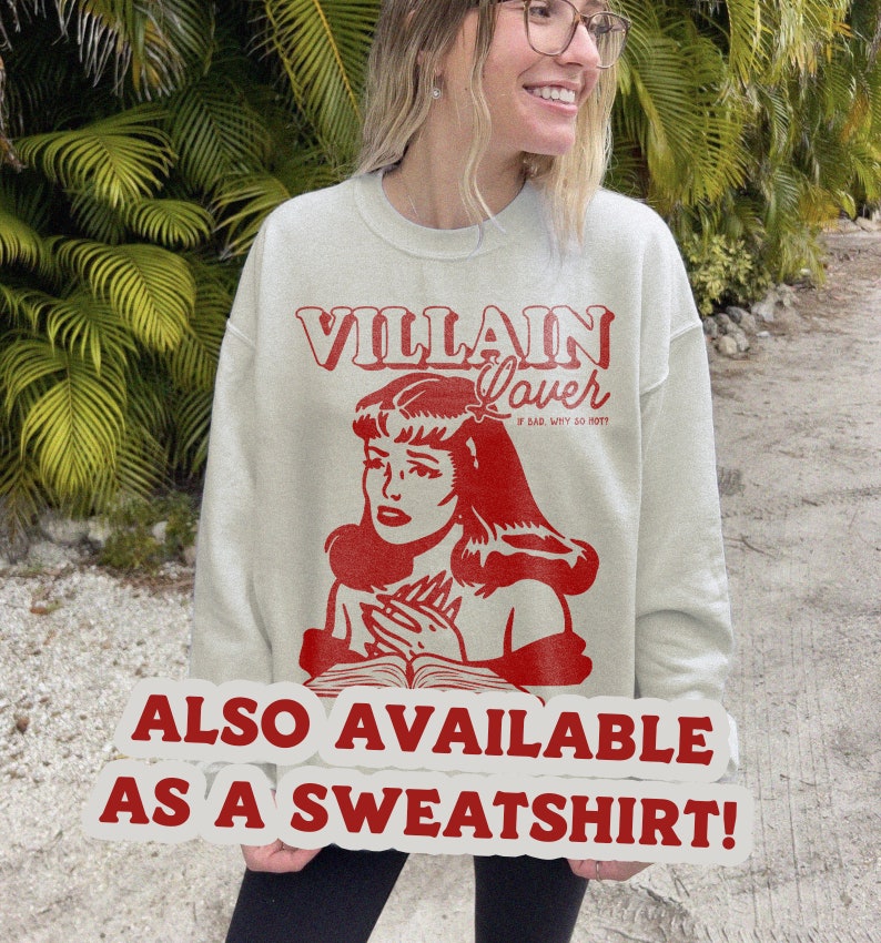 Pode incluir: Uma camisa de moletom cinza claro com uma estampa gr&aacute;fica vermelha de uma mulher com o texto "VILLAIN Lover IF BAD, WHY SO HOT?" e o texto "ALSO AVAILABLE AS A SWEATSHIRT!" em um adesivo branco.