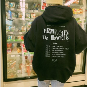 Peut inclure: Un sweat &agrave; capuche noir avec du texte blanc qui dit "Enemies to Lovers Live". Le sweat &agrave; capuche liste &eacute;galement plusieurs dates et titres de chansons. La personne porte un bonnet beige et un jean bleu clair.