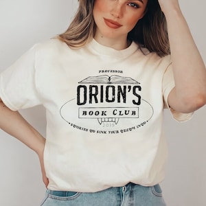 K&ouml;nnte beinhalten: Cremefarbenes T-Shirt mit einem schwarzen Vintage-Design mit der Aufschrift "Professor Orion's Book Club". Das Design zeigt ein offenes Buch, Rei&szlig;z&auml;hne und das Jahr 2019. Der Schriftzug des Shirts enth&auml;lt auch den Satz "Stories to sink your teeth into."