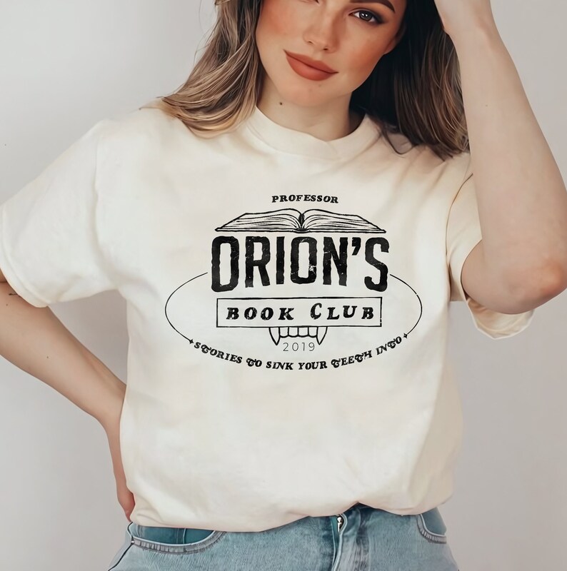 K&ouml;nnte beinhalten: Cremefarbenes T-Shirt mit einem schwarzen Vintage-Design mit der Aufschrift "Professor Orion's Book Club". Das Design zeigt ein offenes Buch, Rei&szlig;z&auml;hne und das Jahr 2019. Der Schriftzug des Shirts enth&auml;lt auch den Satz "Stories to sink your teeth into."