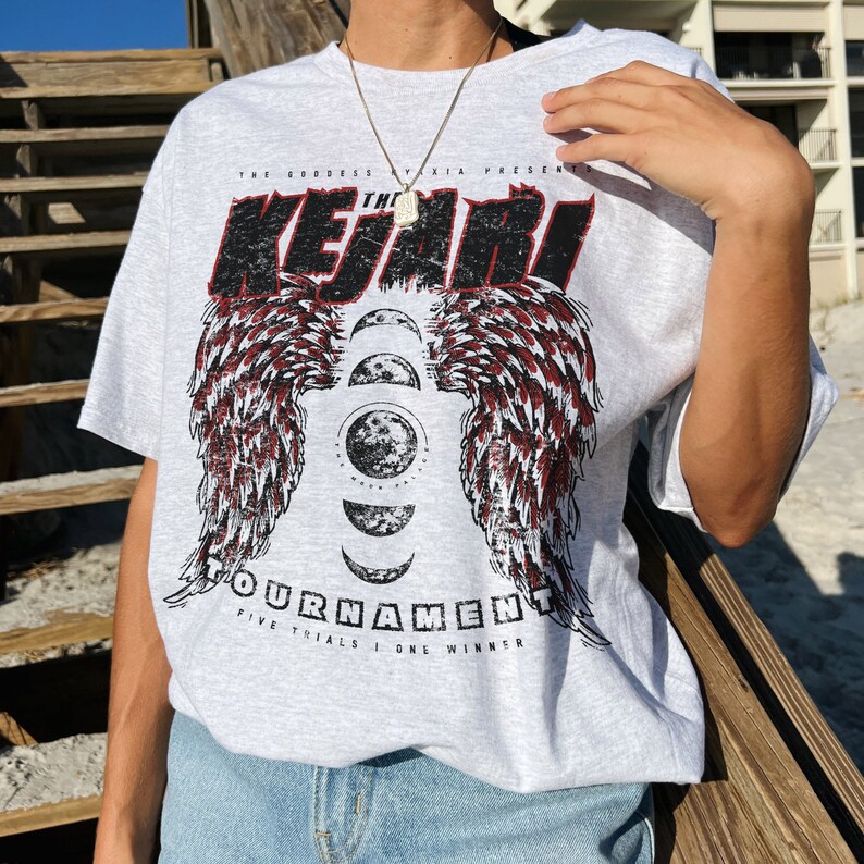 May include: Gray t-shirt with a graphic print of red wings and a moon with the text "The Kejari Tournament Five Trials | One Winner".