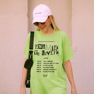 May include: A neon green t-shirt with black text that reads "BOOK ADDIOTS PRESENTS ENEMIES TO LOVERS LIVE" and a list of dates and events. The dates are February 12, 14, 18, 21, 24, and 25. The events are "ONLY ONE BED", "WHO DID THIS TO YOU?", "FATED MATES", "SLOWEST BURN", "FORCED PROXIMITY", and "I'LL KILL FOR YOU".