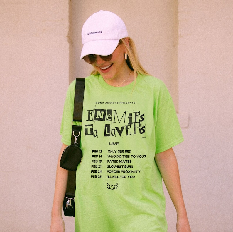 May include: A neon green t-shirt with black text that reads "BOOK ADDIOTS PRESENTS ENEMIES TO LOVERS LIVE" and a list of dates and events. The dates are February 12, 14, 18, 21, 24, and 25. The events are "ONLY ONE BED", "WHO DID THIS TO YOU?", "FATED MATES", "SLOWEST BURN", "FORCED PROXIMITY", and "I'LL KILL FOR YOU".