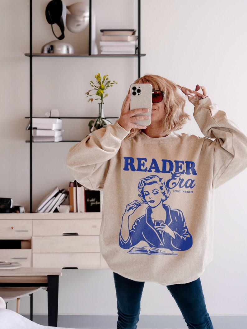 May include: A beige sweatshirt with a blue graphic of a woman reading a book and drinking from a glass. The text on the sweatshirt reads "Reader Era I can't I'm booked".