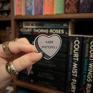 May include: A heart-shaped sticker with the text "KADE MITCHELL" in a black font. The sticker is held in front of a bookshelf filled with books. The books have titles such as "Court of Thorns and Roses" and "Court of Mist and Fury".