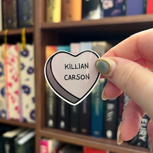 May include: A heart-shaped sticker with the text "KILLIAN CARSON" in a serif font. The sticker has a light gray background with a white border and a dark gray outline. The sticker is held in front of a bookshelf filled with books.