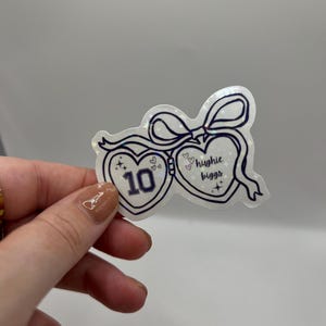 May include: A heart-shaped sticker with a bow and the words "Hughie Biggs" and the number "10" inside. The sticker is white with a holographic sheen and dark blue outlines.
