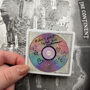 May include: A holographic sticker featuring a CD design with the text "Ridoc's quest Squad jams" and various doodles. The sticker has a square border and is held against a map background. The CD design includes hearts, stars, and sun illustrations.
