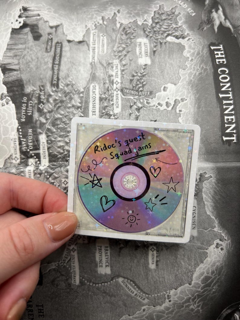 May include: A holographic sticker featuring a CD design with the text "Ridoc's quest Squad jams" and various doodles. The sticker has a square border and is held against a map background. The CD design includes hearts, stars, and sun illustrations.
