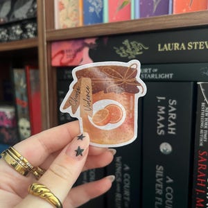 Op de afbeelding: Een sticker met een pot sinaasappelmarmelade met de tekst "Choose Violence". De pot heeft een bruine deksel en een etiket met de woorden "Choose Violence". Twee sinaasappelschijfjes zijn zichtbaar. De sticker wordt voor een boekenplank gehouden.