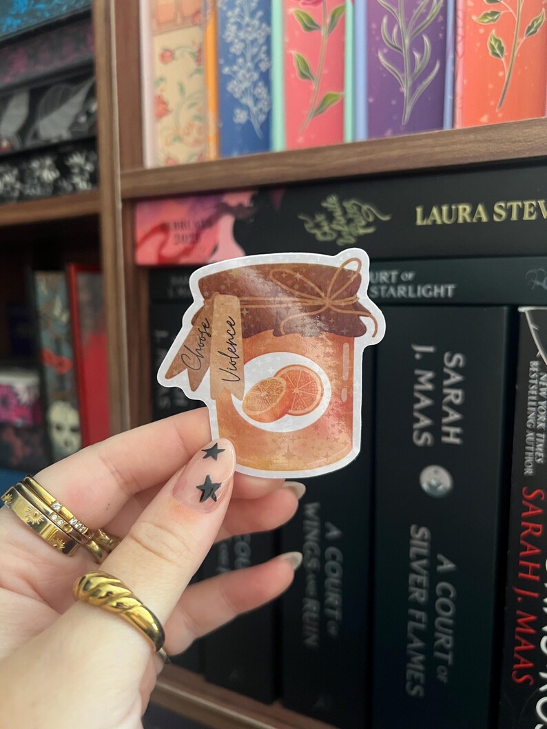 Op de afbeelding: Een sticker met een pot sinaasappelmarmelade met de tekst "Choose Violence". De pot heeft een bruine deksel en een etiket met de woorden "Choose Violence". Twee sinaasappelschijfjes zijn zichtbaar. De sticker wordt voor een boekenplank gehouden.